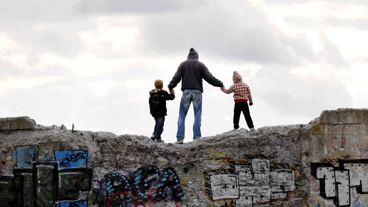 Ein Mann und zwei Kindern stehen Hand in Hand und bei bewölktem Himmel am 11.10.2009 in München auf einem mit Graffitis besprühten Mauerstück auf der Nordheide (Panzerwiese).