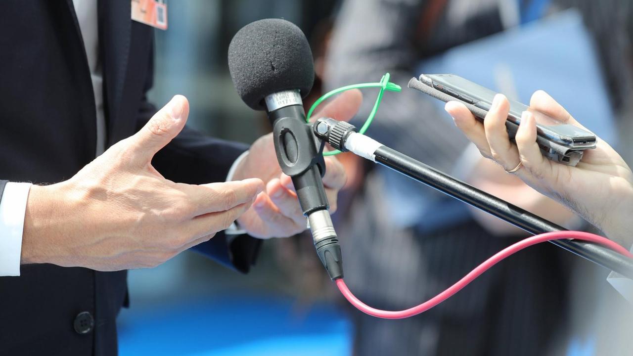 Ein Mikrofon, ein Smartphone und Hände sind in einer Nahaufnahme zu sehen. Ein Mikrofon, ein Smartphone und Hände sind in einer Nahaufnahme zu sehen.