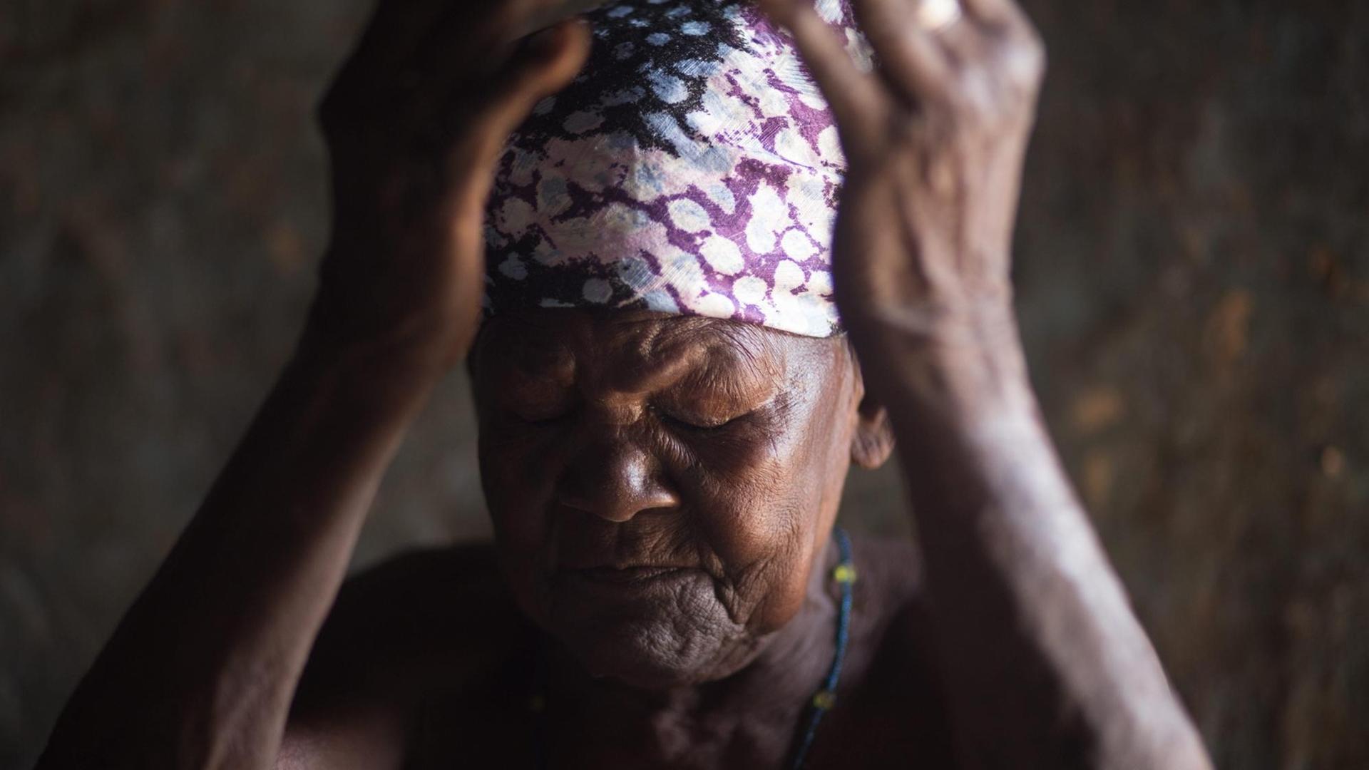 Eine ältere afrikanische Frau, mit einem bunten Kopftuch, hält mit geschlossenen Augen die Hände vor das Gesicht. Eine ältere afrikanische Frau, mit einem bunten Kopftuch, hält mit geschlossenen Augen die Hände vor das Gesicht.