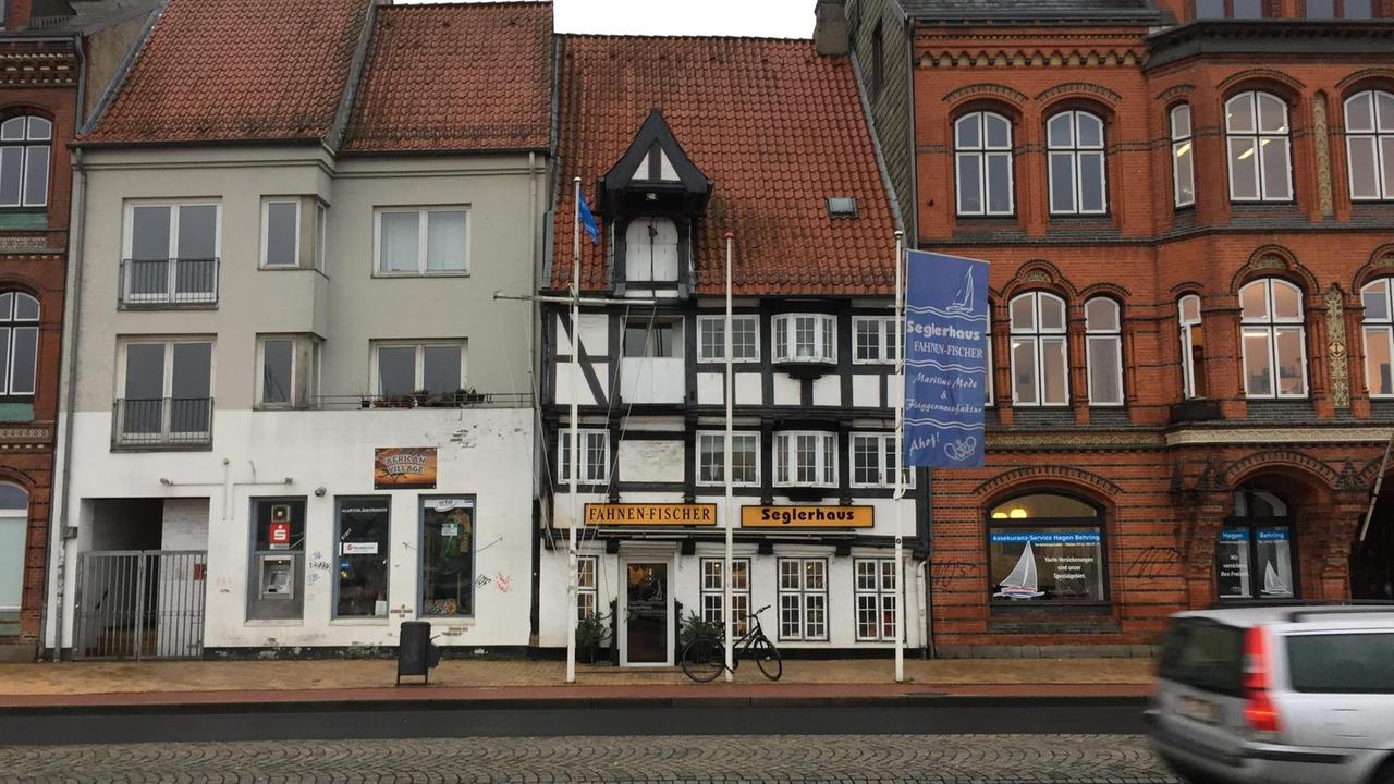 In der Mitte steht ein Fachwerkhaus, mit dem Namen "Fahnen Fischer" in schwarzen Buchstaben auf gelbem Banner über der Tür.  Umgeben von anderen Häusern in Flensburg.