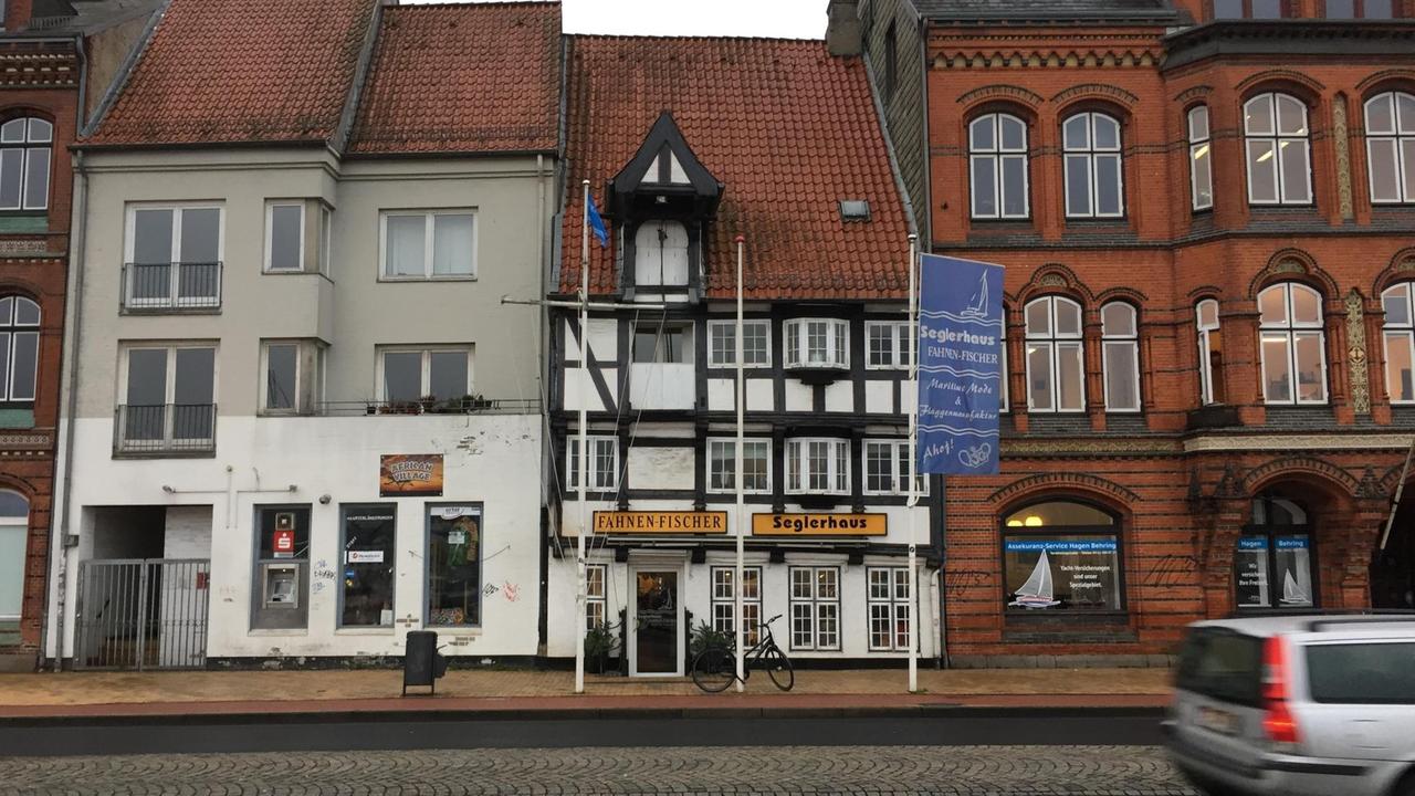 In der Mitte steht ein Fachwerkhaus, mit dem Namen "Fahnen Fischer" in schwarzen Buchstaben auf gelbem Banner über der Tür. Umgeben von anderen Häusern in Flensburg. In der Mitte steht ein Fachwerkhaus, mit dem Namen "Fahnen Fischer" in schwarzen Buchstaben auf gelbem Banner über der Tür. Umgeben von anderen Häusern in Flensburg.