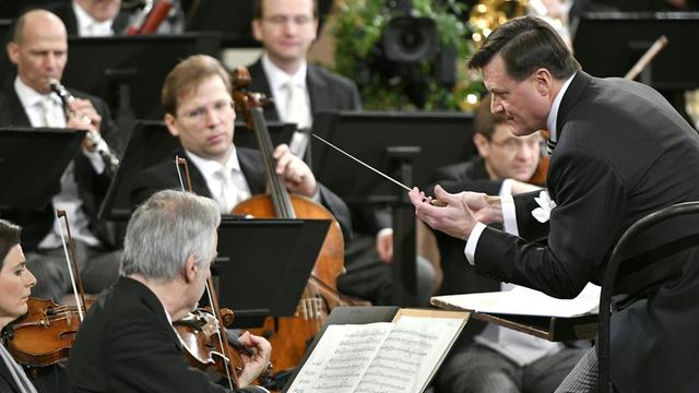 Ein Mann in schwarzem Frack und Taktstock in der rechten Hand ist von seiner linken Seiten aus zu sehen, wie er gebückt vor einem Orchester steht.