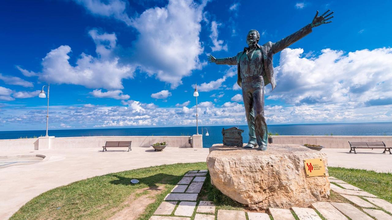 Eine Statue von Domenico Modugno an der Meeresküste vor strahlend blauem Himmel. Modugno breitet seine Arme weit aus und lacht.