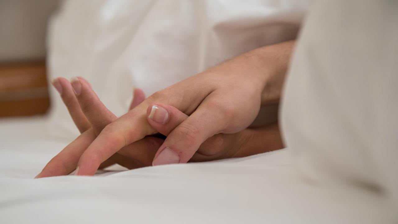 Die Hände eines Mannes und einer Frau sind zwischen zerknitterten Laken in einem Bett zu sehen.