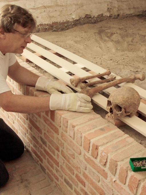 Andreas Ströbl kniet am Boden und zeigt Schädel und Knochen die auf einer Bank liegen.