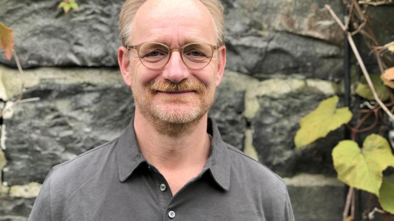 Ein lächelnder, halbkahler Mann mit Brille und rötlichem Schnurrbart vor einer efeubewachsenen Mauer. Ein lächelnder, halbkahler Mann mit Brille und rötlichem Schnurrbart vor einer efeubewachsenen Mauer.
