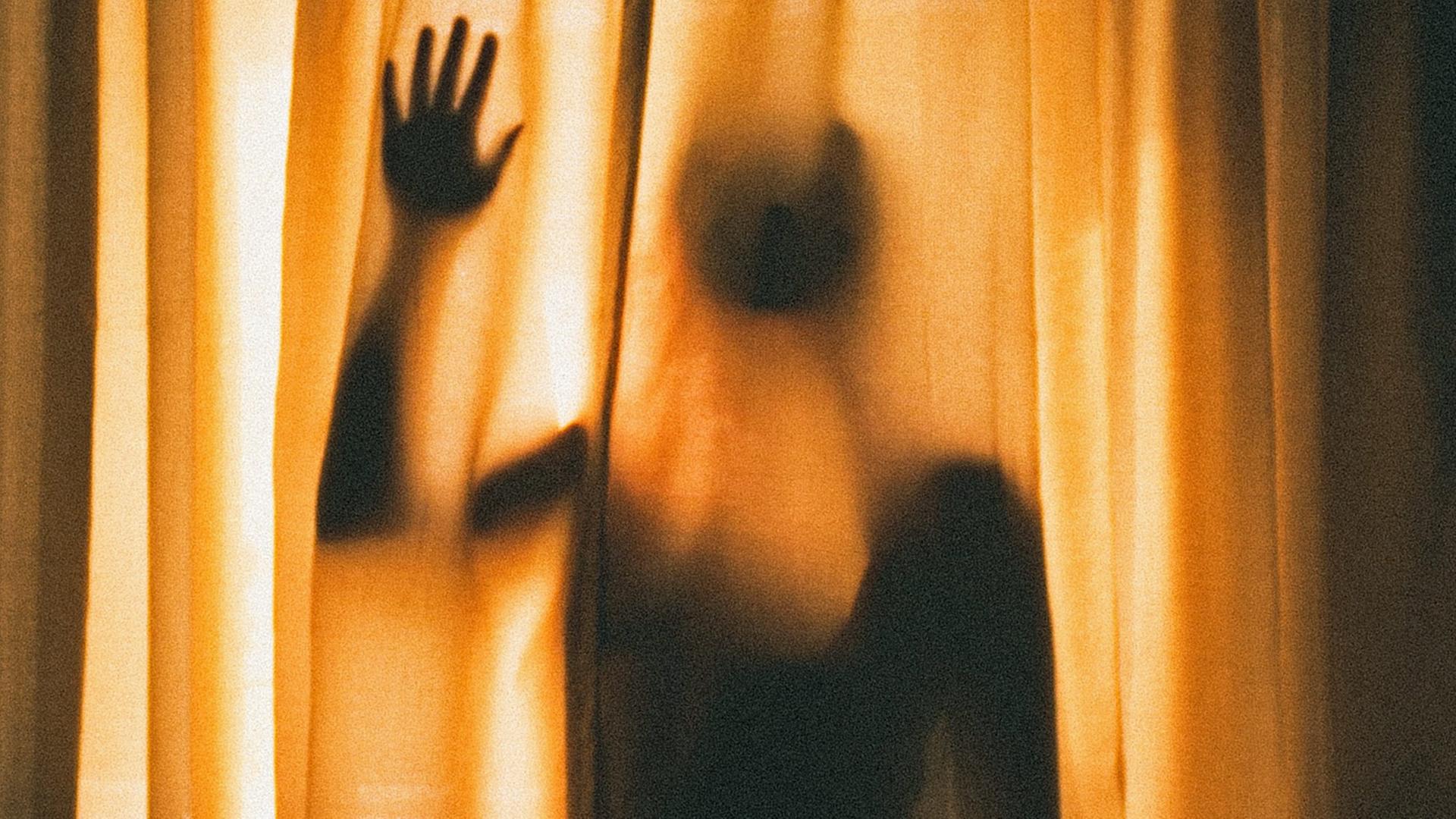 Eine Person steht im Gegenlicht hinter einem transparenten Vorhang und berührt diesen Eine Person steht im Gegenlicht hinter einem transparenten Vorhang und berührt diesen