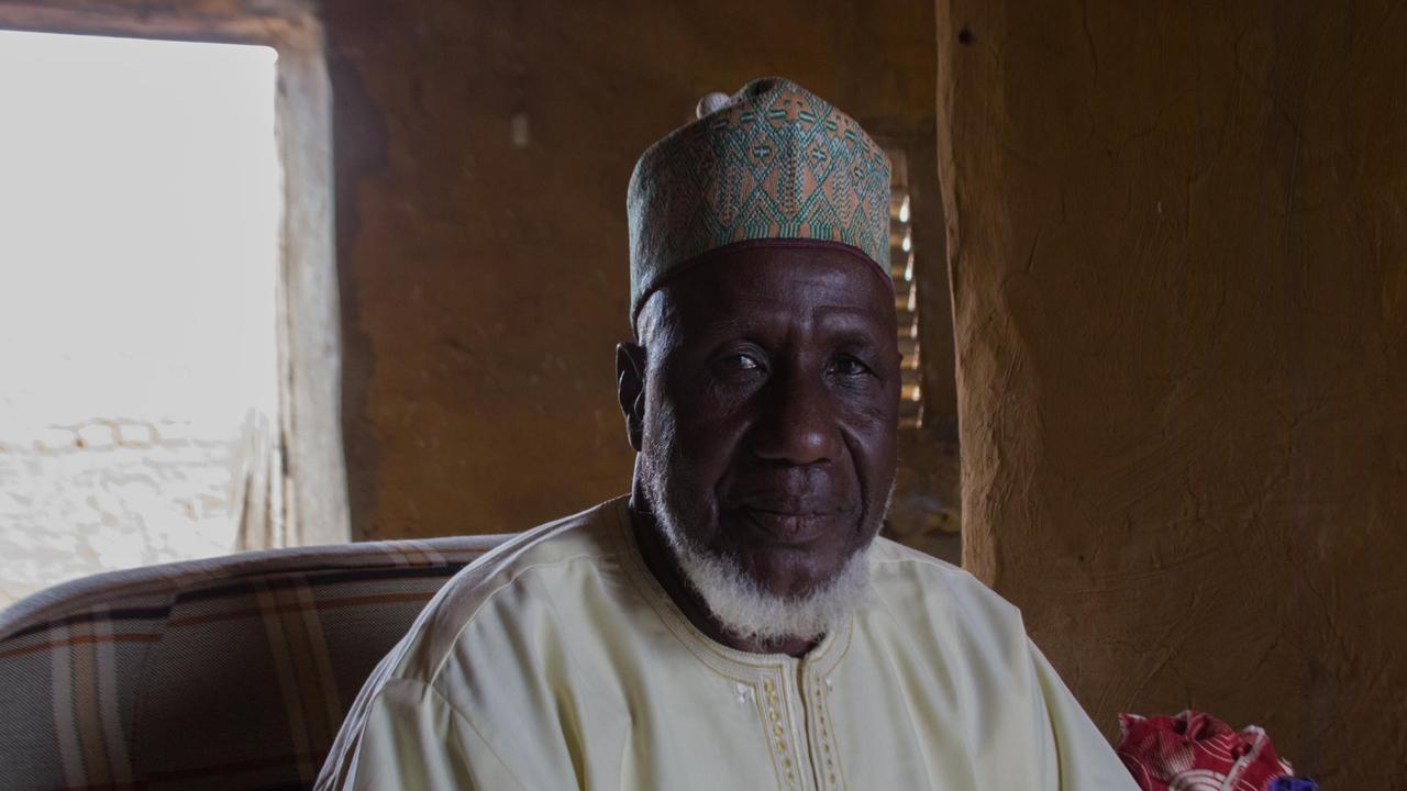 Ein älterer afrikanischer Mann mit Kappe und weißem Gewand sitzt auf einem Stuhl. Ein älterer afrikanischer Mann mit Kappe und weißem Gewand sitzt auf einem Stuhl.