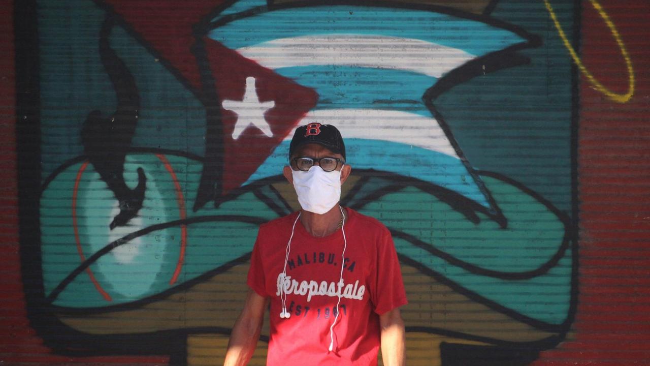 Ein Mann mit Mundschutz steht in Havanna vor einem Wandbild mit der kubanischen Flagge. 