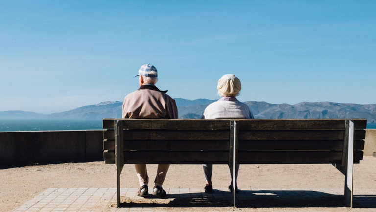 Zwei ältere Menschen sitzen auf einer Parkbank und gucken auf ein Bergpanorama Zwei ältere Menschen sitzen auf einer Parkbank und gucken auf ein Bergpanorama
