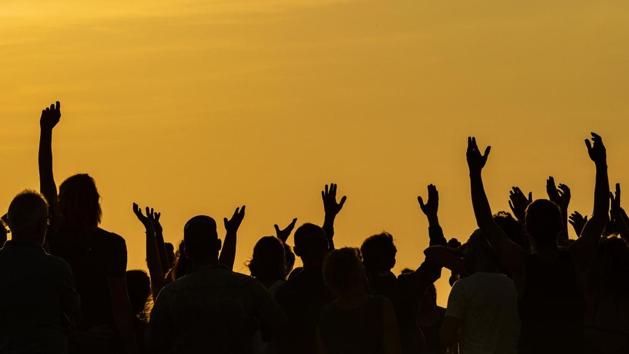 Eine Menschengruppe hebt im Schattenriss die Arme vor einer glutroten Abendstimmung. Eine Menschengruppe hebt im Schattenriss die Arme vor einer glutroten Abendstimmung.