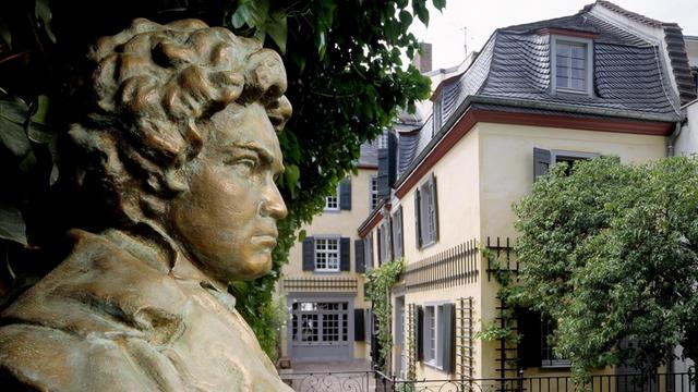 Außenansicht eines cremefarbenen Hauses mit Dachschindeln und Fensterläden in Dunkelgrau. Im Vordergrund die efeuumrankte Steinbüste von Ludwig van Beethovens Kopf.
