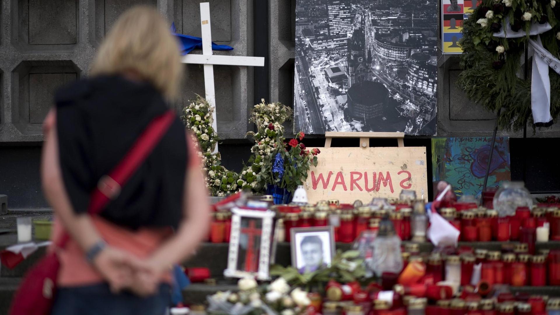 Eine Frau steht vor Kerzen und Blumen und einem Schild mit der Aufschrift "Warum" am Berliner Breitscheidplatz.