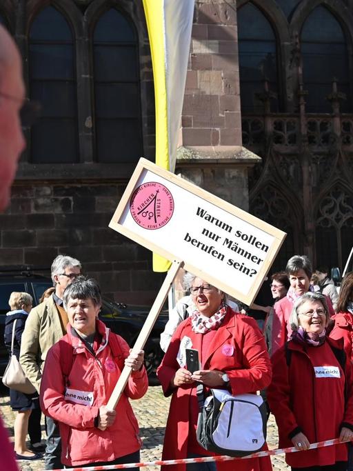 Priester laufen vor dem Münster an Teilnehmern einer Protestaktion der Initiative Maria 2.0 der Katholische Frauengemeinschaft in der Erzdiözese Freiburg vorbei. Auf einem Plakat steht "Warum sollen nur Männer berufen sein?".