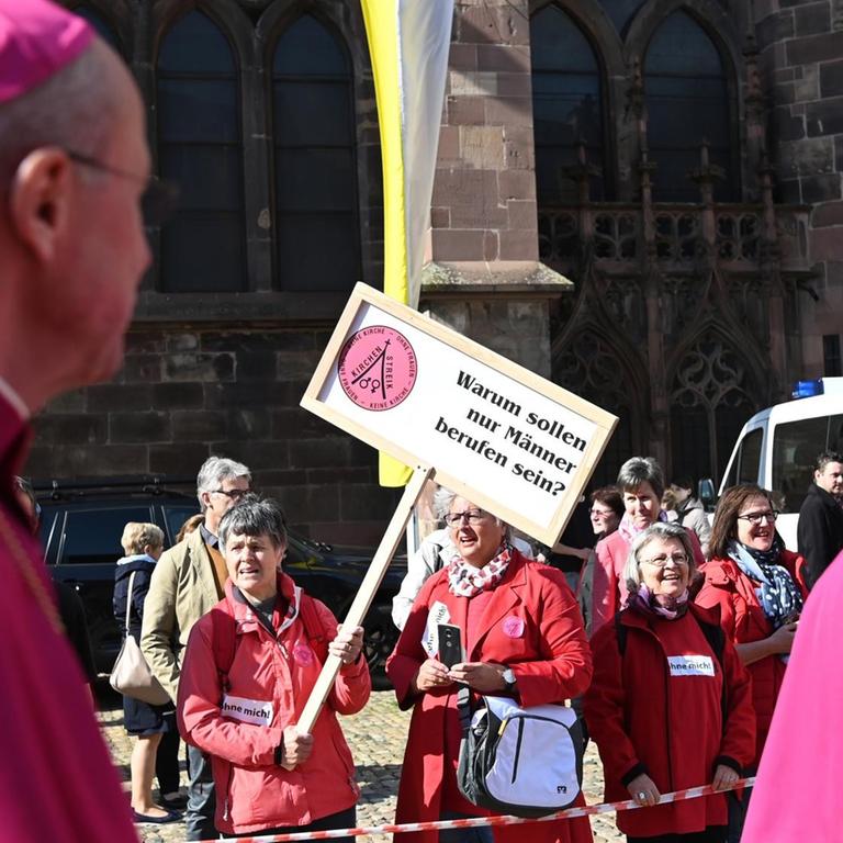 Priester laufen vor dem Münster an Teilnehmern einer Protestaktion der Initiative Maria 2.0 der Katholische Frauengemeinschaft in der Erzdiözese Freiburg vorbei. Auf einem Plakat steht "Warum sollen nur Männer berufen sein?".