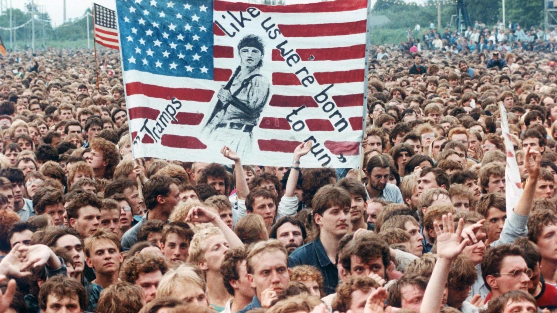 Das Foto vom Publikum beim Bruce-Springsteen-Konzert am 19.07.1988 in Ost-Berlin zeigt begeisterte, junge Männer. Einige Menschen halten ein selbstgemaltes Plakat hoch, darauf eine US-Flagge und die Aufschrift "We Tramps we're born to run" - "Wir Tramps, wir sind geboren, um zu rennen." Das Foto vom Publikum beim Bruce-Springsteen-Konzert am 19.07.1988 in Ost-Berlin zeigt begeisterte, junge Männer. Einige Menschen halten ein selbstgemaltes Plakat hoch, darauf eine US-Flagge und die Aufschrift "We Tramps we're born to run" - "Wir Tramps, wir sind geboren, um zu rennen."