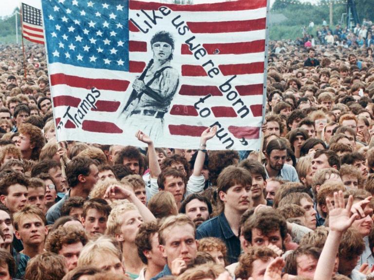 Das Foto vom Publikum beim Bruce-Springsteen-Konzert am 19.07.1988 in Ost-Berlin zeigt begeisterte, junge Männer. Einige Menschen halten ein selbstgemaltes Plakat hoch, darauf eine US-Flagge und die Aufschrift "We Tramps we're born to run" - "Wir Tramps, wir sind geboren, um zu rennen."