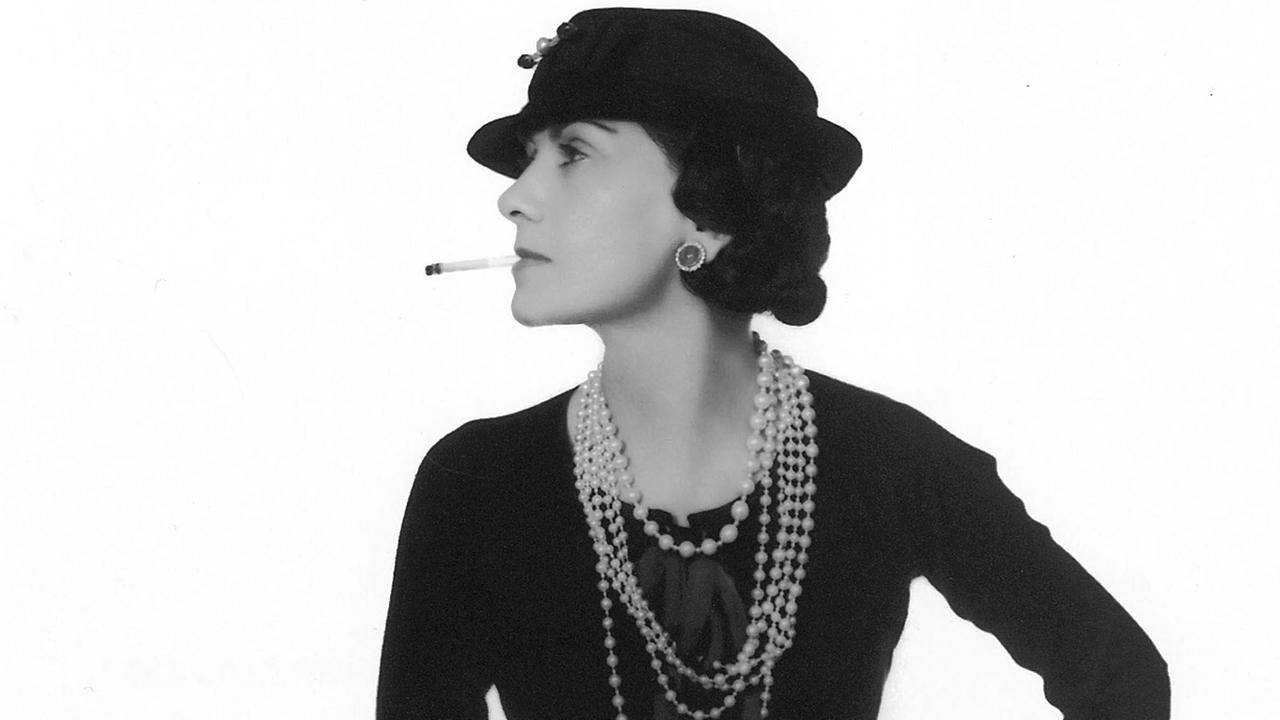 Coco Chanel mit Zigarette im Mund, Perlenketten um den Hals und Hut auf dem Kopf, nach links schauend, in Schwarz-Weiß