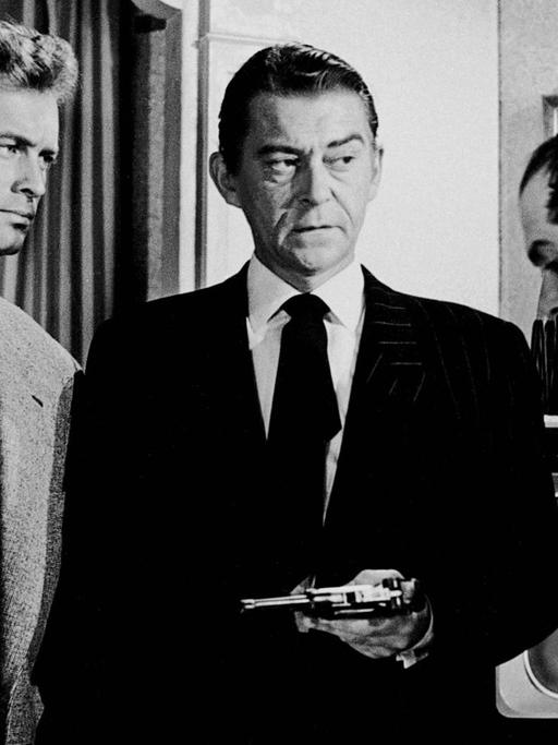 Szene aus dem Film "Rififi" (1955) von Jules Dassin. Drei Männer stehen zusammen und schmieden einen Plan. Der in der Mitte hält eine Waffe in der Hand.