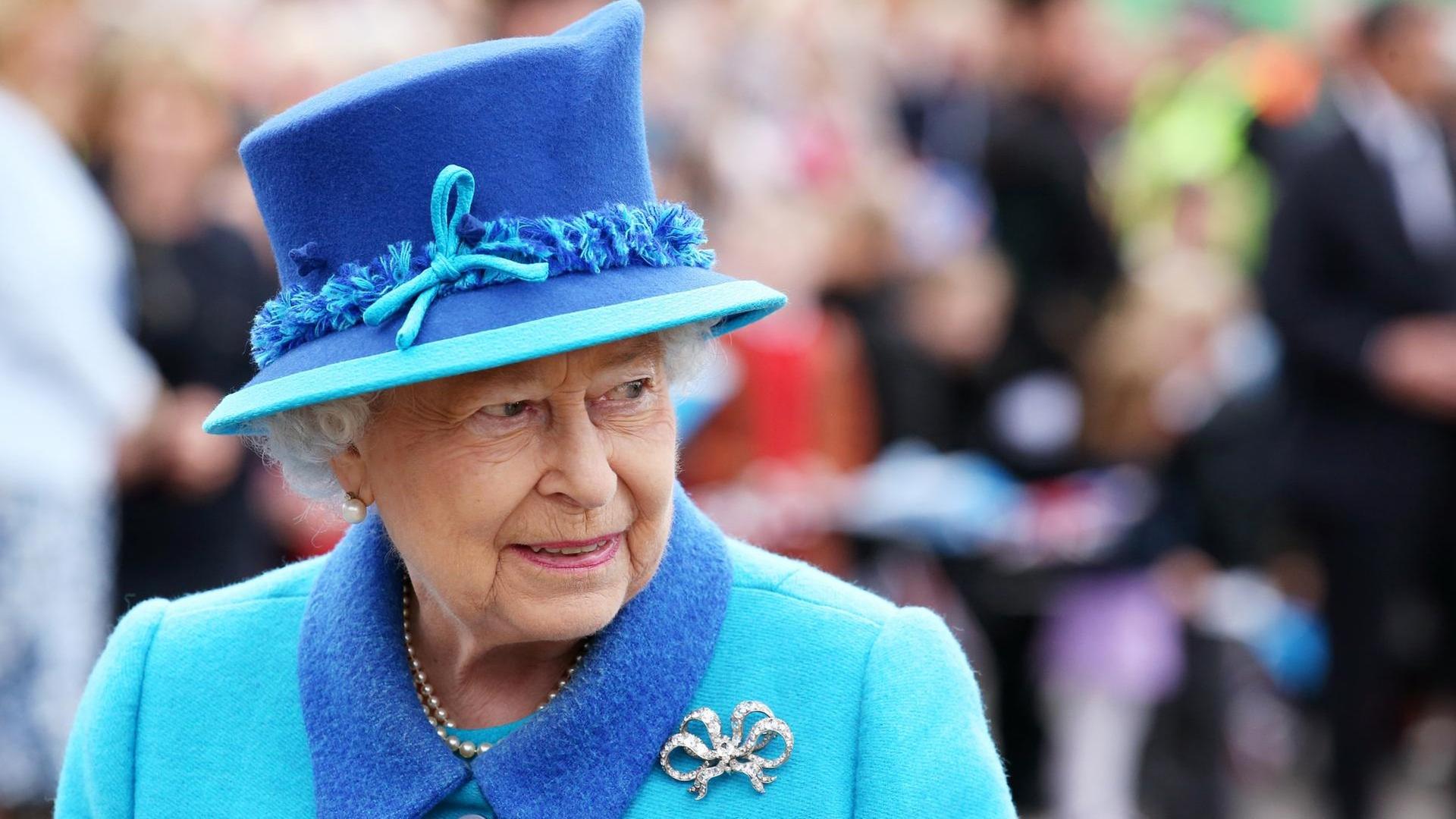 Die Queen steht im hellblauen Kostüm mit dunkelblauem Hut und silberner Brosche im Freien vor einer Menschenmenge, die im Hintergrund unscharf zu erkennen ist. Die Queen steht im hellblauen Kostüm mit dunkelblauem Hut und silberner Brosche im Freien vor einer Menschenmenge, die im Hintergrund unscharf zu erkennen ist.
