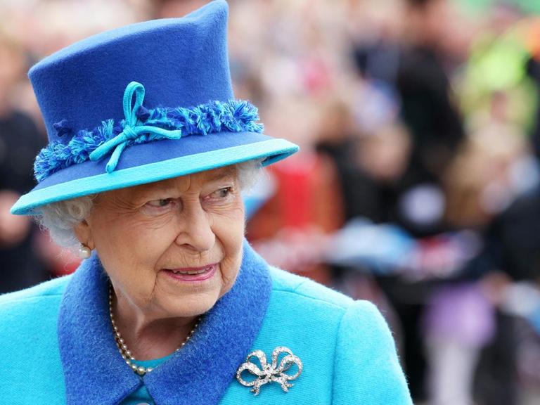 Die Queen steht im hellblauen Kostüm mit dunkelblauem Hut und silberner Brosche im Freien vor einer Menschenmenge, die im Hintergrund unscharf zu erkennen ist.