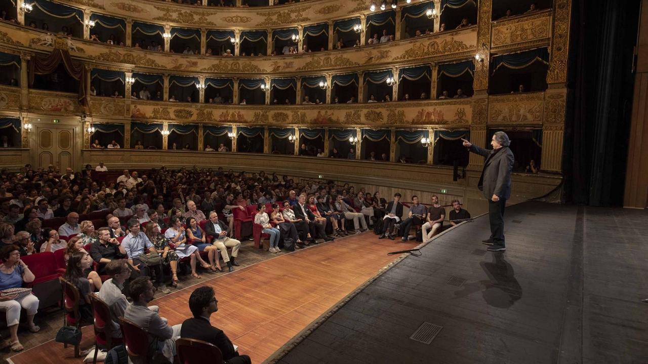 Riccardo Muti steht mit ausgestrecktem Arm auf der Bühne des Teatro Alighieri in Ravenna, im Zuschauerraum lauschen zahlreiche Besucher seinen Ausführungen