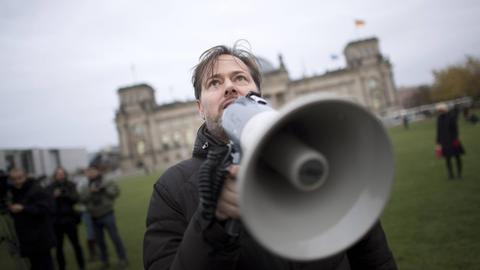 Milo Rau steht mit Megafon auf der Wiese vor dem Reichstag.