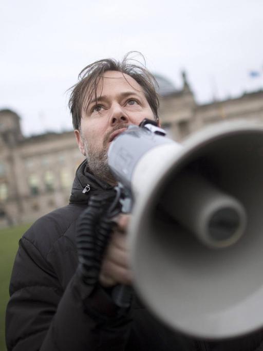 Milo Rau steht mit Megafon auf der Wiese vor dem Reichstag.