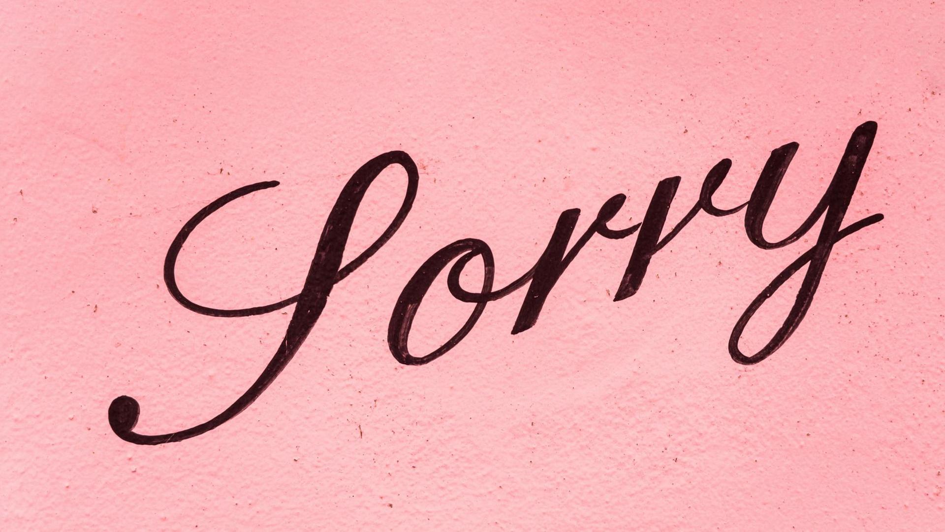 Auf einer rosafarbenen Hauswand steht in schwarzer Schreibschrift „Sorry”