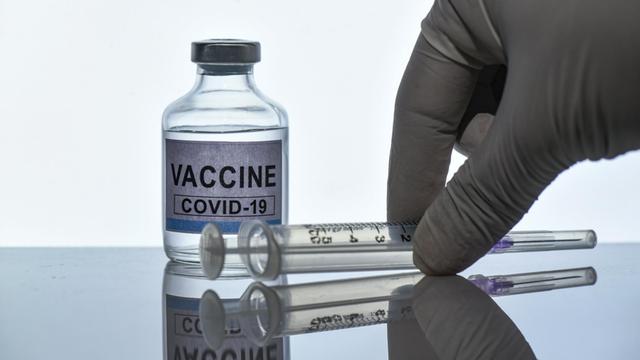 Symbolbild eines Glasfläschchens, auf dem "Vaccine Covid-19" steht, dazu eine behandschuhte Hand und eine Spritze.