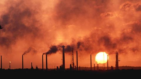 Schornsteine einer Erdölraffinerie stoßen schwarzen Rauch vor einem roten Himmel bei Sonnenuntergang aus.