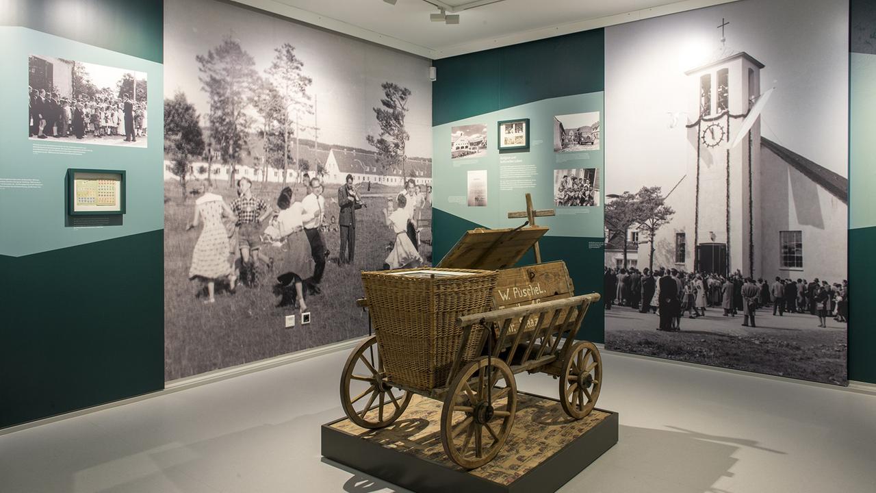 Blick in die Ausstellung des Erinnerungsorts Badehaus im ehemaligen DP-Lager Föhrenwald und heutigen Waldram in Oberbayern: ein Leiterwagen, umgeben von Bildwänden, auf denen unter anderem eine Kirche mit Menschen davor zu sehen ist. Blick in die Ausstellung des Erinnerungsorts Badehaus im ehemaligen DP-Lager Föhrenwald und heutigen Waldram in Oberbayern: ein Leiterwagen, umgeben von Bildwänden, auf denen unter anderem eine Kirche mit Menschen davor zu sehen ist.