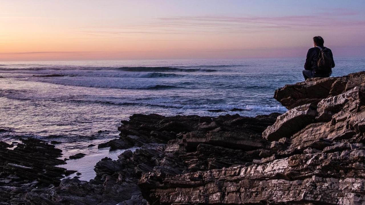 Hinteransicht eines Mannes, der auf einer Felsküste sitzt und aufs weite Meer hinaus blickt. Hinteransicht eines Mannes, der auf einer Felsküste sitzt und aufs weite Meer hinaus blickt.