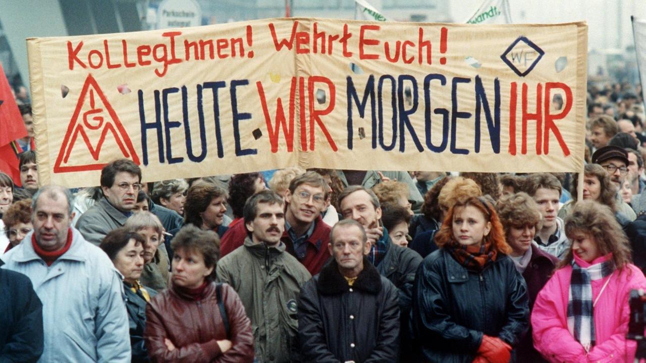 Demonstranten tragen ein Transparent, auf dem steht: "Liebe KollegInnen! Wehrt Euch! Heute wir, morgen ihr"