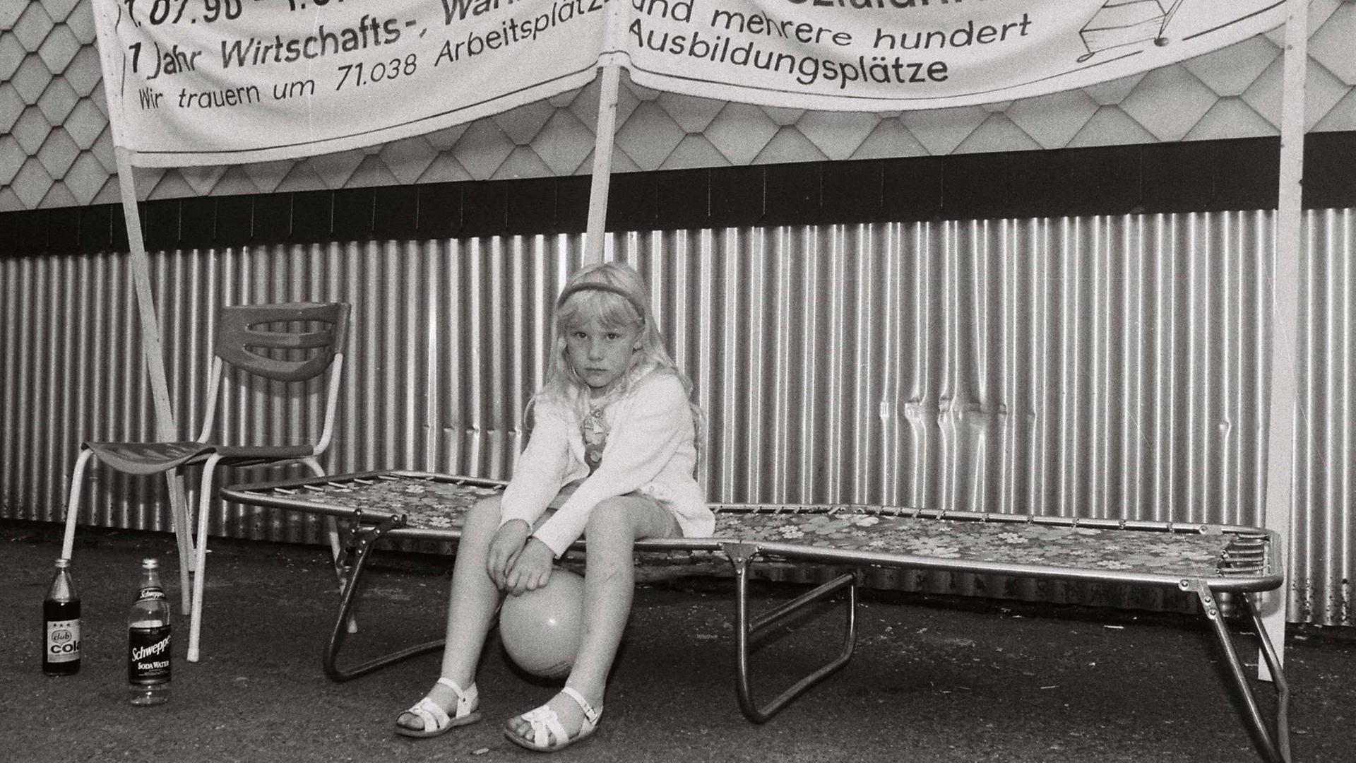 Ein kleines Mädchen sitzt anläßlich der Besetzung der Treuhand-Niederlassung in Suhl/Thüringen am 01.07.1991 mit einem Luftballon zwischen den Beinen auf einer Camping-Liege. Über Ihr auf einem Transparent steht "1.07.90-1-07.91 - 1 Jahr Wirtschafts-, Währungs- und Sozialunion. Wir Trauern um 71.038 Arbeitsplätze und mehrere hundert Ausbildungsplätze." Ein kleines Mädchen sitzt anläßlich der Besetzung der Treuhand-Niederlassung in Suhl/Thüringen am 01.07.1991 mit einem Luftballon zwischen den Beinen auf einer Camping-Liege. Über Ihr auf einem Transparent steht "1.07.90-1-07.91 - 1 Jahr Wirtschafts-, Währungs- und Sozialunion. Wir Trauern um 71.038 Arbeitsplätze und mehrere hundert Ausbildungsplätze."