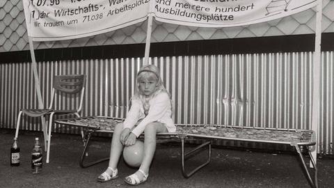 Ein kleines Mädchen sitzt anläßlich der Besetzung der Treuhand-Niederlassung in Suhl/Thüringen am 01.07.1991 mit einem Luftballon zwischen den Beinen auf einer Camping-Liege. Über Ihr auf einem Transparent steht "1.07.90-1-07.91 - 1 Jahr Wirtschafts-, Währungs- und Sozialunion. Wir Trauern um 71.038 Arbeitsplätze und mehrere hundert Ausbildungsplätze."