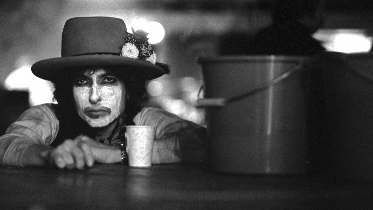 Der Musiker Bob Dylan, die Hände auf einer Tischplatte, mit weiß geschminktem Gesicht und dunklem Hut sowie mit einem Kaffeebecher vor und einem Putzeimer neben sich. Der Musiker Bob Dylan, die Hände auf einer Tischplatte, mit weiß geschminktem Gesicht und dunklem Hut sowie mit einem Kaffeebecher vor und einem Putzeimer neben sich.