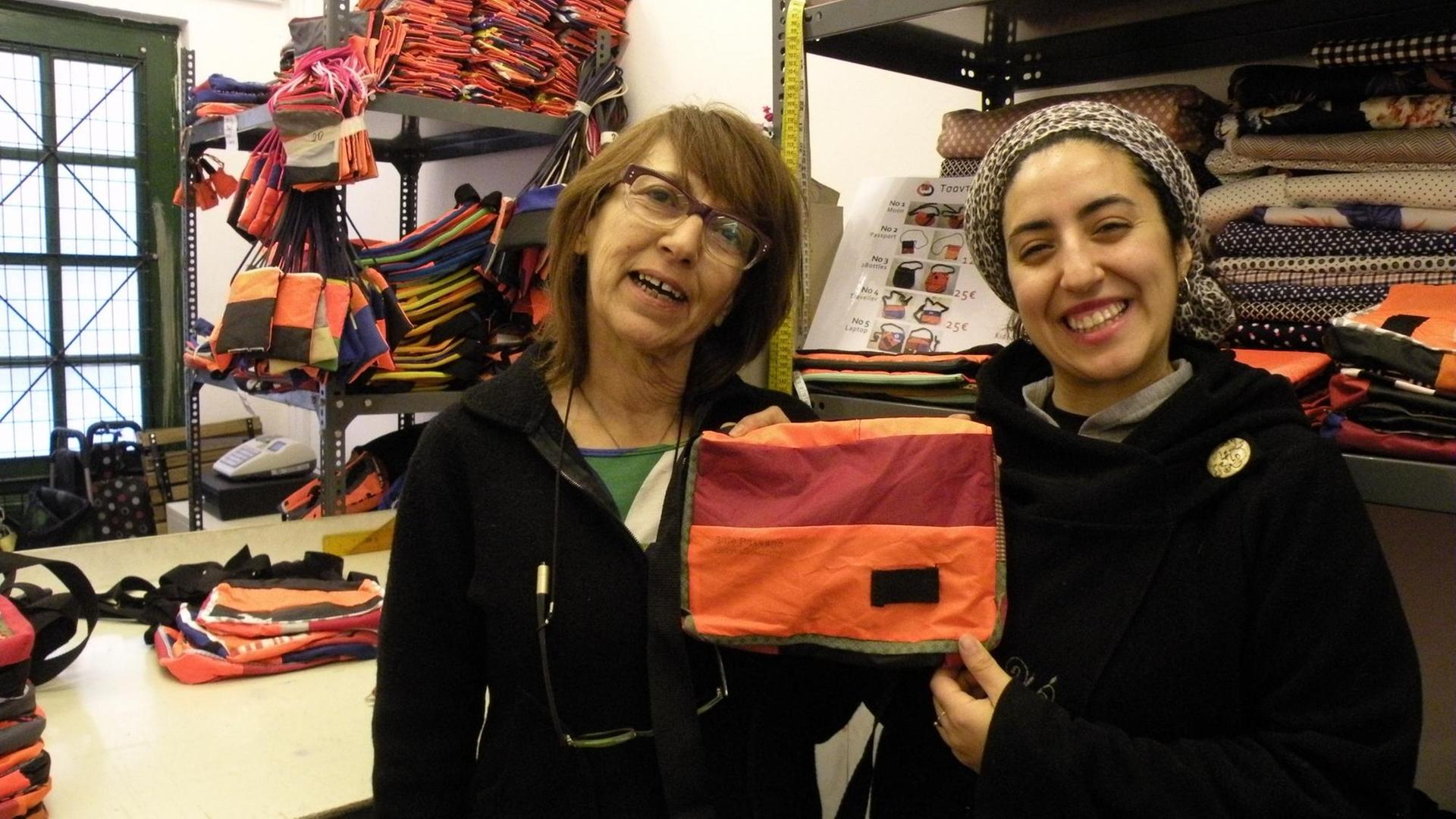 Zwei Frauen: Matina(links) und Sofia (rechts) halten eine handgemachte Tasche hoch. Im Hintergrund das Material, alte Rettungswesten gestapelt. Zwei Frauen: Matina(links) und Sofia (rechts) halten eine handgemachte Tasche hoch. Im Hintergrund das Material, alte Rettungswesten gestapelt.