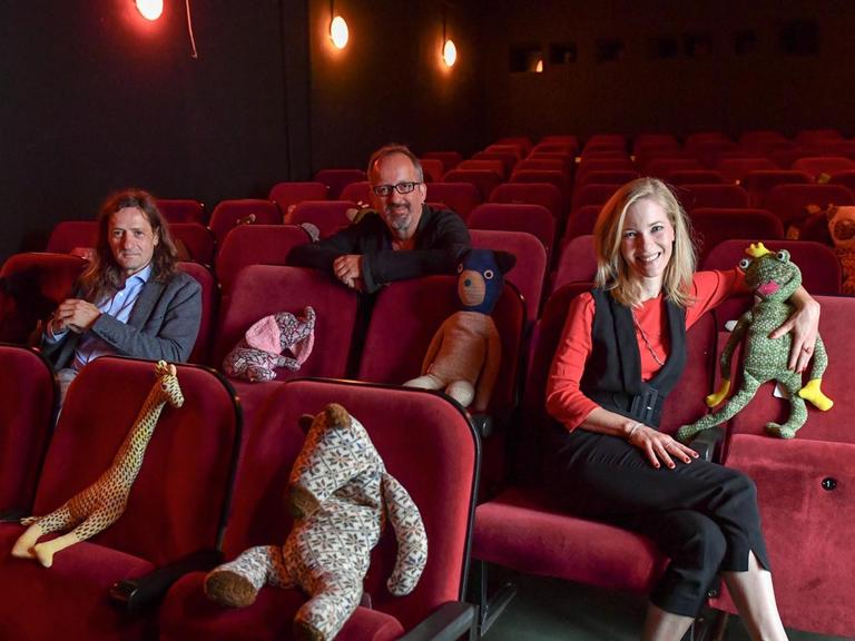 Sascha Grunow, Christos Acrivulis und Martina Klier (v.l.n.r.) sitzen im Kinosaal in roten Sesseln. Auf Sesseln dazwischen haben sie Kuscheltiere platziert.