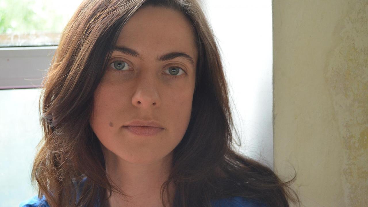 Kateryna Mishchenko steht mit dem Rücken zu einem Fenster, sie trägt halblanges Haar und eine blaue Bluse und schaut ernst in die Kamera. Kateryna Mishchenko steht mit dem Rücken zu einem Fenster, sie trägt halblanges Haar und eine blaue Bluse und schaut ernst in die Kamera.