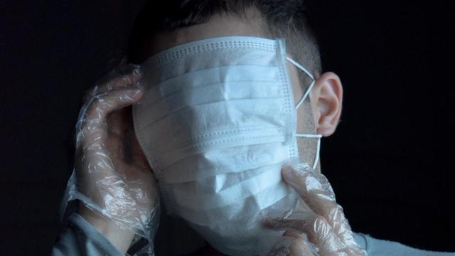 Ein Mann mit Plastikhandschuhen trägt zwei Atemschutzmasken und verdeckt mit der einen seine Augen.