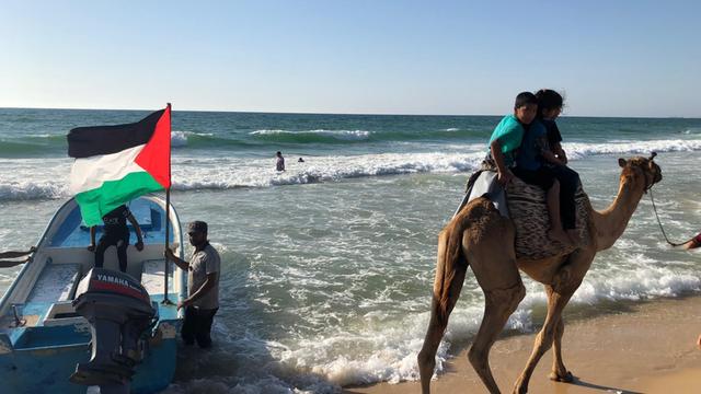 Menschen am Strand und zwei die auf einem Kamel reiten, zusehen ein Fischerboot mit palästinensischer Flagge