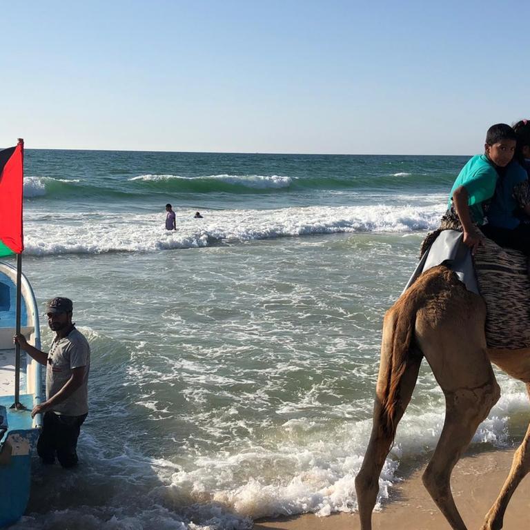 Menschen am Strand und zwei die auf einem Kamel reiten, zusehen ein Fischerboot mit palästinensischer Flagge
