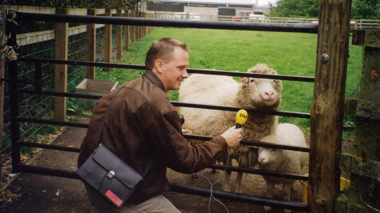Der DLF Reporter Michael Lange hält dem Klonschaf Dolly ein Mikrofon unter die Schnauze. Dolly steht mit ihren Lämmern auf einer Weide und guckt den Journalisten durch das Gatter an. Der DLF Reporter Michael Lange hält dem Klonschaf Dolly ein Mikrofon unter die Schnauze. Dolly steht mit ihren Lämmern auf einer Weide und guckt den Journalisten durch das Gatter an.