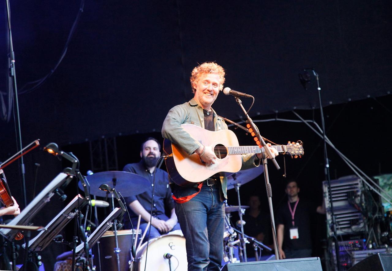 Der Musiker Glen Hansard steht lächelnd, die Gitarre um den Hals auf einer Bühne und spielt 