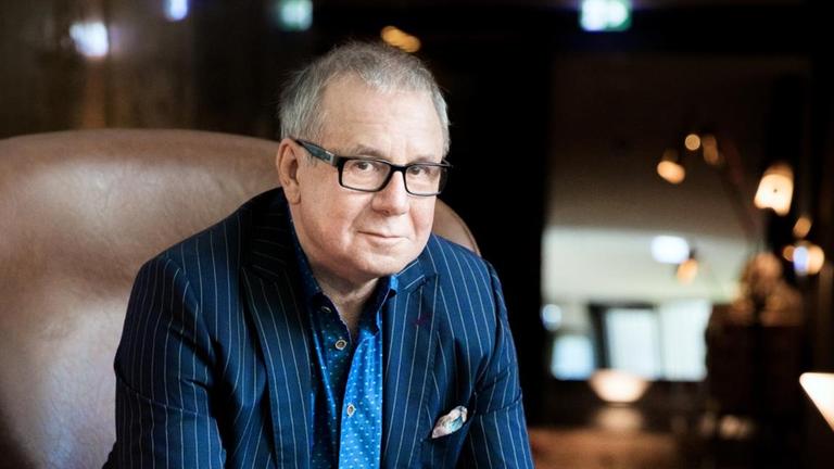 Der Schauspieler Jocahim Król sitzt in einem brauen Designer-Sessel und schaut in die Kamera. Er trägt einen blauen Nadelstreifen-Anzug und eine Brille. Der Schauspieler Jocahim Król sitzt in einem brauen Designer-Sessel und schaut in die Kamera. Er trägt einen blauen Nadelstreifen-Anzug und eine Brille.