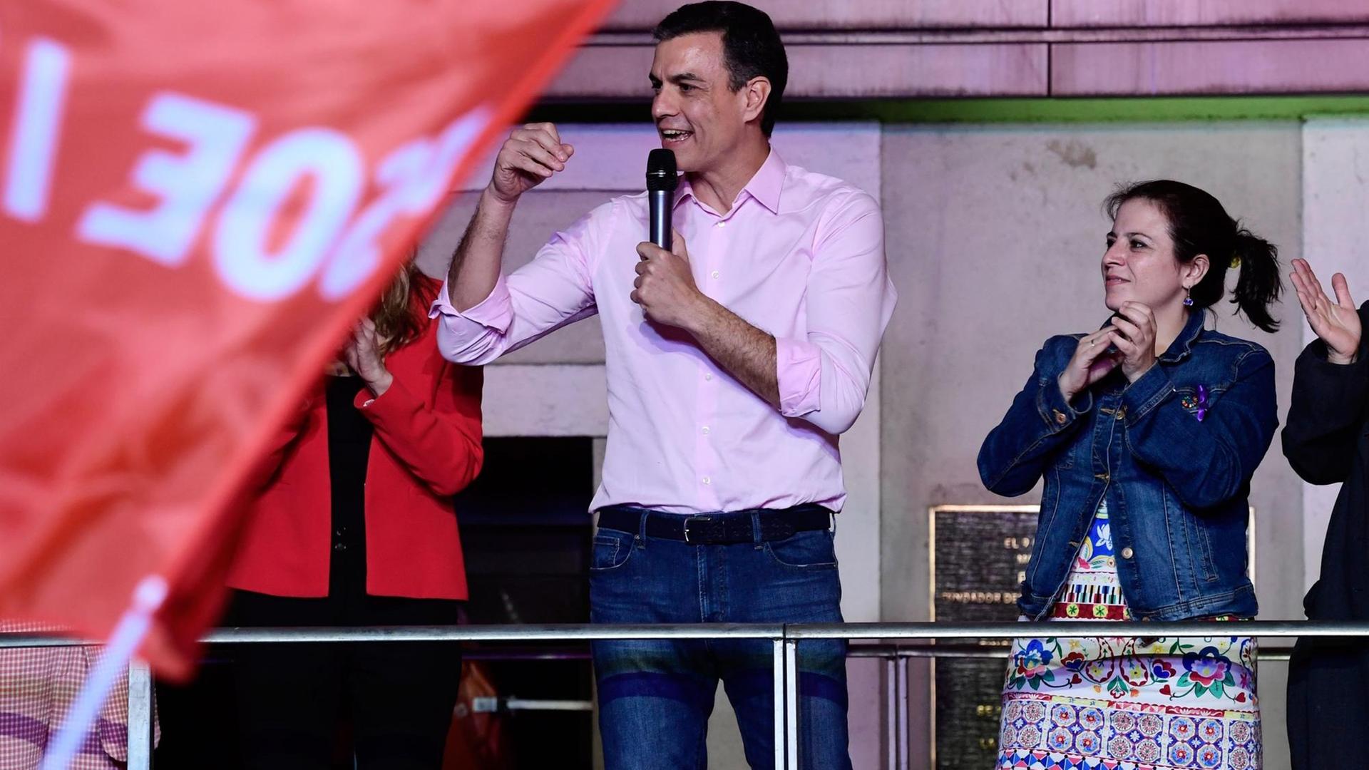 Ministepräsident Sanchez aus Spanien steht auf einer Bühne und feiert seinen Wahlsieg. Links die Fahne seiner sozialistischen Partei.