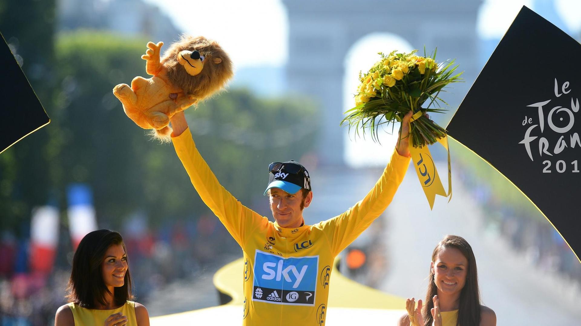 Bradley Wiggins trägt ein gelbes Trikot und reckt in Siegerpose beide Arme in die Höhe. In der rechten Hand hat er einen Plüsch-Löwen, in der linken einen Strauß mit gelben Blumen. Bradley Wiggins trägt ein gelbes Trikot und reckt in Siegerpose beide Arme in die Höhe. In der rechten Hand hat er einen Plüsch-Löwen, in der linken einen Strauß mit gelben Blumen.