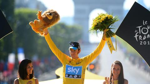 Bradley Wiggins trägt ein gelbes Trikot und reckt in Siegerpose beide Arme in die Höhe. In der rechten Hand hat er einen Plüsch-Löwen, in der linken einen Strauß mit gelben Blumen.