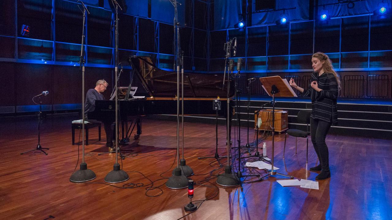 In einem holzgetäfelten Konzertsaal stehen viele Mikrofone, rechts im Bild ist eine junge Sängerin zu sehen, links ein Pianist am schwarzen Flügel. In einem holzgetäfelten Konzertsaal stehen viele Mikrofone, rechts im Bild ist eine junge Sängerin zu sehen, links ein Pianist am schwarzen Flügel.